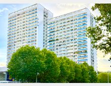 Hochhaus mit weißer Fassade und zahlreichen Balkonen in einem modernen architektonischen Stil, umgeben von grünen Baumkronen im Vordergrund unter einem leicht bewölkten Himmel.