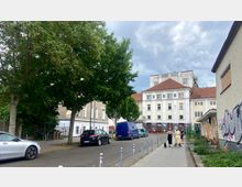Straßenszene in einer urbanen Umgebung mit Bäumen, parkenden Autos und Gehweg; im Hintergrund ein mehrstöckiges Gebäude mit heller Fassade und rotem Dach, auf dem große Buchstaben "B B B" zu sehen sind. Die Umgebung wirkt teilweise mit Graffiti versehen und leicht verlassen.