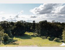 Weite Wiesenfläche mit vereinzelten Sträuchern und einer dichten Baumreihe im Hintergrund unter einem teilweise bewölkten Himmel. Am rechten Bildrand ist ein weißes, zeltähnliches Objekt zu sehen.