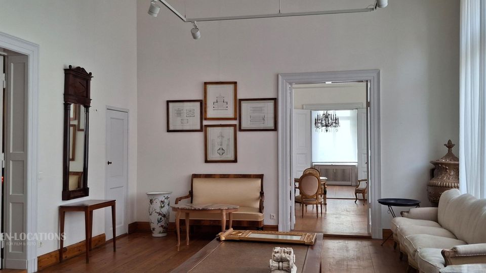 Heller Wohnraum im klassischen Stil mit weißen Wänden, Holzfußboden und großen Fenstern mit Vorhängen. Zu sehen sind Sitzmöbel, ein Wandspiegel, gerahmte Zeichnungen sowie breite Türdurchgänge in angrenzende Räume, darunter ein Raum mit Kronleuchter.