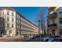 Straßenszene mit mehrstöckigen, historischen Wohngebäuden mit verzierten Fassaden, Balkonen und roten Dächern. Geparkte Autos säumen die Straße, während kahle Bäume und ein blauer Himmel den städtischen Charakter unterstreichen.