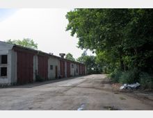 Ein Industriegebäude mit grauer Fassade und mehreren großen, braunen Toren entlang einer asphaltierten Fläche, umgeben von dichter Vegetation und Bäumen auf der rechten Seite. Der Himmel ist hell und offen, ohne sichtbare Wolken.