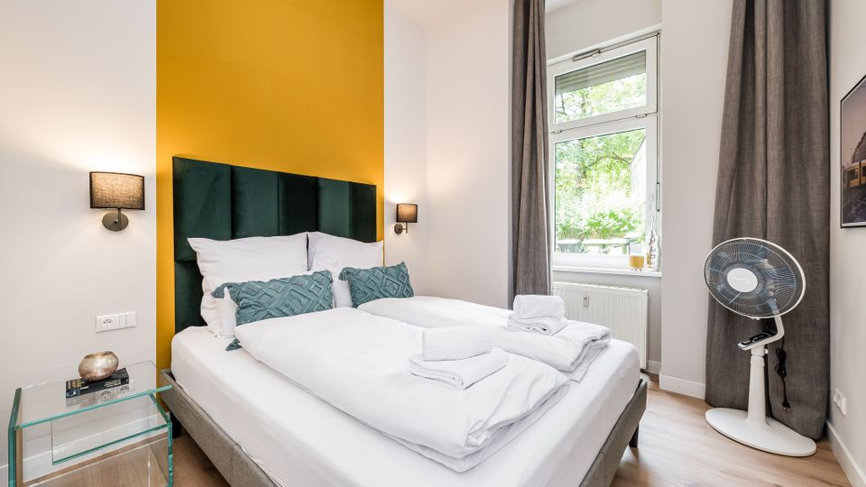 Modernes Schlafzimmer mit Doppelbett, weißer Bettwäsche und dunkelgrün gepolstertem Kopfteil vor einer gelben Akzentwand, flankiert von zwei Wandlampen. Rechts ein großes Fenster mit grauen Vorhängen, Heizkörper darunter, ein Standventilator sowie ein kleiner Glas-Beistelltisch auf hellem Holzfußboden.