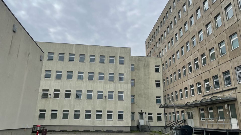 Innenhof eines mehrstöckigen Beton-Gebäudekomplexes mit vielen rechteckigen Fenstern und einer gepflasterten, teils rissigen Hoffläche. Rechts ist ein Eingang mit Vordach und Rampe zu sehen, im Hof stehen ein großer Müllcontainer und ein roter Hydrant.