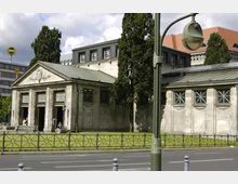Historisches Gebäude mit einer Fassade im Stil eines klassischen Tempels, flankiert von großen Fenstern mit Kreuzmustern; im Hintergrund moderne Bürogebäude und hohe Bäume. Im Vordergrund führt eine Straße mit Laternen und Geländern vorbei.