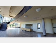 Innenansicht eines leerstehenden Gebäudes mit einer geschwungenen Treppe links, großflächigen Fensterfronten, einer Rezeption in der Mitte und abblätternder Farbe an der Decke. Rechts befindet sich eine weiß geflieste Säule sowie eine Garderobe mit leeren Kleiderstangen.