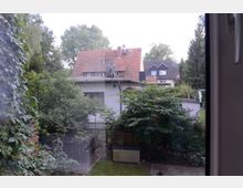 Blick aus einem Fenster auf einen kleinen Garten mit Bäumen und Sträuchern, dahinter ein weißes Flachdachgebäude und ein rotes Haus mit Satteldach. Im Hintergrund sind weitere Häuser und hohe Bäume zu sehen.