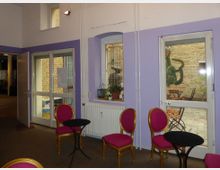 Ein kleiner Innenraum mit lila Wänden, großen Fenstern und Glastüren, die auf einen gepflasterten Innenhof führen. Im Raum stehen Tische mit schwarzen Oberflächen und pink gepolsterte Stühle mit goldenen Rahmen, im Innenhof sind weitere Sitzgelegenheiten und eine bemalte Wand zu sehen.