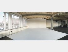 Weitläufiger, industrieller Innenraum mit sichtbarer Betondecke und großen Fenstern, die Tageslicht einlassen. Der Raum hat weiße und schwarze Böden, eine freistehende Säule sowie angrenzende Bereiche mit Möbeln und einer kleinen Küchenzeile.