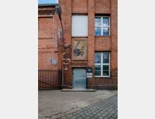 Rote Backsteinfassade eines Industriegebäudes mit großen Fenstern, schmiedeeisernem Zaun links und einer Eingangstür mit Hausnummer „401“. Über der Tür befindet sich ein Gemälde mit einer Arbeiterfigur und einem Werkzeug.