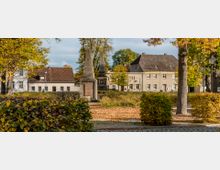 Ein kleiner Park im Herbst mit buntem Laub auf dem Boden, umgeben von mehreren Wohnhäusern. Im Vordergrund stehen zwei steinerne Obelisken, und Büsche sowie Bäume mit gelb-orangenen Blättern säumen die Szene.