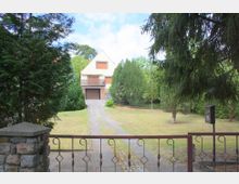 Ein Einfamilienhaus mit Satteldach, hellgelber Fassade und geschlossenen Rollläden, steht am Ende einer langen Auffahrt. Der Garten ist von Bäumen und Sträuchern umgeben, und im Vordergrund befindet sich ein schmiedeeisernes Tor mit einer kleinen Mauer.