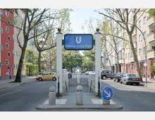 Eingang zur U-Bahn-Station „Viktoria-Luise-Platz“ in Berlin, umgeben von mehrstöckigen Wohngebäuden, Bäumen und geparkten Autos entlang der Straße. Das blaue Stationsschild ist an weißen Pfeilern befestigt, und eine Treppe führt hinunter zur unterirdischen Station.