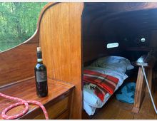 Blick in die Schlafkajüte eines kleinen Holzbootes mit dunkel getäfeltem, gebogenem Innenraum und schmalen Fenstern. Im Vordergrund steht auf einer Holzbank eine Weinflasche neben einem Seil; in der Kajüte liegen Bettzeug und Decken, seitlich ist ein kleiner Klapptisch zu sehen.