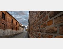 Schmale, kopfsteingepflasterte Gasse mit historischen roten Ziegelmauern auf beiden Seiten, links ein Gebäude mit teils verputzter Fassade und kleinen Fenstern, rechts eine hohe Backsteinmauer mit einer Laterne. Im Hintergrund sind weitere Gebäude und ein bewölkter Himmel sichtbar.