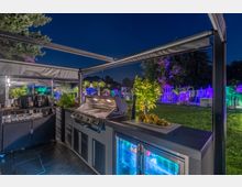 Eine moderne Außenküche bei Nacht mit einem Grill, Kühlschränken und Arbeitsflächen, umgeben von einem beleuchteten Garten mit Rasen, Palmen und bunten Lichteffekten. Im Hintergrund sind Bäume, Büsche und Gartenmöbel sichtbar.