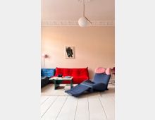 Ein Wohnzimmer mit hellen Wänden, einer dekorativen Stuckverzierung entlang der Decke und einer modernen Hängelampe. Im Raum stehen ein rotes Sofa, ein blaues Sofa und ein blauer Liegesessel mit rosa Kopfstützen, dazu ein kleiner Couchtisch auf einem hellen Teppichboden.