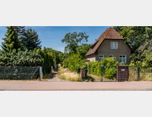 Wohnhaus mit Satteldach und beigem Putz, umgeben von dichtem Grün und Bäumen, hinter einem Drahtzaun mit einer gemauerten Säule am Eingang. Rechts daneben verläuft ein schmaler, unbefestigter Weg entlang von Hecken und Bäumen.