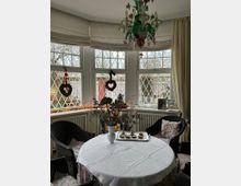 Ein Wintergarten mit halbkreisförmigen Fenstern im klassischen Stil, geschmückt mit Gittersprossen und Kränzen. Im Vordergrund steht ein runder Tisch mit weißer Tischdecke, umgeben von Korbstühlen; darüber ein kunstvoller Lüster.