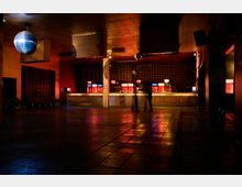 Leerer Nachtclub mit einem großen, beleuchteten Tresen im Hintergrund, einer spiegelnden Discokugel an der Decke und gedimmter Beleuchtung, die den Raum in rot-orange Töne taucht. Der Boden besteht aus großen Fliesen, und vereinzelte Sitzgelegenheiten befinden sich an den Seitenwänden.