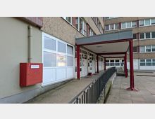 Eingangsbereich eines mehrstöckigen Wohn- oder Verwaltungsgebäudes mit überdachter Rampe und Metallgeländer, weißen Fenster- und Türfronten sowie einem roten Brandschutzkasten an der Außenwand. Der Zugang liegt in einem gepflasterten Innenhof mit sichtbaren Rohrleitungen und Stützen unter dem Vordach.