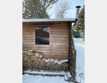 Ein kleiner Holzschuppen mit einer verschneiten Holzfassade und einem Blechkamin auf dem Dach. Vor dem Schuppen liegt gestapeltes Brennholz, während im Hintergrund Bäume und ein schneebedeckter Garten zu sehen sind.
