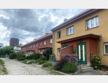 Reihenhäuser in einer Wohnsiedlung mit farbigen Fassaden in Gelb und Rot, Vorgärten mit Pflanzen und einem gepflasterten Gehweg davor; im Hintergrund ist ein Backsteinturm mit grünem Dach zu sehen, unter einem bewölkten Himmel.