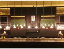 Eine Bar mit glänzender Holztheke, von hängenden Lampen beleuchtet. Im Hintergrund stehen Regale mit Gläsern, Dekorationen und kleinen Pflanzen vor einer Wand mit grünem Fliesenmuster. Ein Schild weist auf zusätzliche Bars auf den Emporen hin.