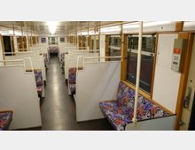 Innenraum eines älteren Zugwaggons mit Sitzbänken in einem offenen Abteil-Layout. Die Wände sind hell, die Sitzpolster haben bunte Muster, und entlang der Fenster verlaufen Handläufe aus Metall.