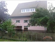 Zweistöckiges Haus mit rosafarbener Fassade, braunem Ziegeldach und kleinen weißen Fenstern. Ein einfacher Holzzaun umgibt den Garten, der mit Bäumen und Sträuchern bepflanzt ist.