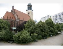 Backsteinkirche mit einem hohen Kirchturm und roten Ziegeldach, umgeben von dichtem Grün mit Bäumen und Sträuchern. Im Vordergrund ist ein gepflasterter Platz mit Parkbänken und vereinzelten Personen zu sehen, während moderne Wohngebäude im Hintergrund stehen.