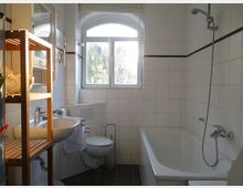 Badezimmer mit weiß gefliesten Wänden, einer Badewanne mit Duscharmatur rechts, einem Waschbecken mit Spiegel links und einem Fenster mit Rundbogen in der Mitte. Ein Holzregal steht neben dem Waschbecken, der Boden ist dunkel gefliest.