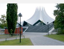 Modernes Kulturgebäude mit einer markanten, weißen, spitzen Dachkonstruktion, die an Zeltspitzen erinnert. Vor dem Gebäude führen breite Treppen hinauf, umgeben von Grünflächen, Bäumen, Laternen und einem roten Kiosk.