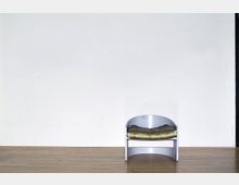 Ein moderner Raum mit hellem Holzboden und einer weißen Wand im Hintergrund. Vor der Wand steht ein einzelner, minimalistischer Metallstuhl mit einem olivgrünen Kissen.