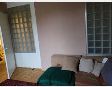 Wohnzimmer mit einer schlichten Einrichtung, darunter ein beiger L-förmiger Sofa mit mehreren Kissen und einer grünen Decke. Links und rechts an der Wand befinden sich große Vorrichtungen aus Glasbausteinen; daneben ist eine offene weiße Tür zu sehen, die in einen angrenzenden Raum führt. Der Boden besteht aus Holzparkett.