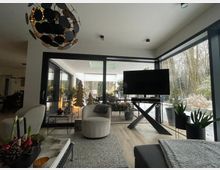 Moderner Wohnbereich mit großer Glasschiebetür und bodentiefen Fenstern, die auf eine überdachte Terrasse und einen verschneiten Garten mit Bäumen hinausgehen. Im Raum stehen ein Sessel auf Teppich, ein Fernseher auf einem X-förmigen Tisch sowie Pflanzen und eine markante Deckenleuchte.