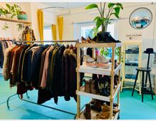 Ein Secondhand-Laden mit hängender Kleidung auf einer Metallstange, einem Regal mit Schuhen und Pflanzen sowie gelben Vorhängen vor großen Fenstern. Im Hintergrund ein runder Wandspiegel und ein Schrank mit Accessoires.