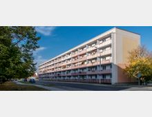 Ein mehrstöckiges Wohngebäude mit pastellrosa und weißen Balkonen entlang der gesamten Fassade, gelegen an einer ruhigen Straße mit Gehweg und Bäumen auf der linken Seite. Im Vordergrund sind parkende Autos zu sehen, und die Szene wird von klarem Himmel und herbstlichen Bäumen ergänzt.