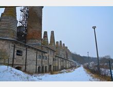 Alte Industrieanlage mit einer Reihe von hohen, zylindrischen Schornsteinen aus Ziegeln, die an ein längliches Gebäude aus Steinmauerwerk angrenzen; umgeben von schneebedecktem Boden und kahlen Bäumen.