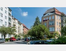 Wohnstraße mit mehrstöckigen Altbauten und roten Ziegeldächern, umgeben von Bäumen und geparkten Autos. Der Himmel ist klar und blau, mit vereinzelten Wolken.