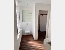 Helles Badezimmer mit weißer Wandgestaltung, einem wandhängenden WC, gläsernen Regalböden in einer Nische und einem Fenster links mit halbtransparentem Rollo. Der Boden besteht aus Holzlaminat, und eine Tür im Hintergrund führt in einen weiteren Raum.