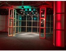 Ein moderner Innenraum mit Holzrahmenkonstruktionen und Glasabtrennungen, beleuchtet durch rote und grüne Lichteffekte. Im Hintergrund sind glatte Wandflächen und dekorative Hängelampen erkennbar, die eine geometrische Anordnung betonen.