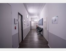 Ein heller, moderner Flur mit grauem Fußboden und weiß gestrichenen Wänden. Auf der rechten Seite befinden sich schwarze Stühle entlang der Wand, links sind mehrere Türen und Wandinstallationen wie ein Desinfektionsspender sichtbar. Auf beiden Seiten hängen Schilder und Pläne.