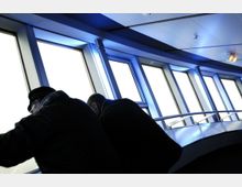 Innenraum einer Aussichtsplattform mit geneigten Fenstern, die Tageslicht hereinlassen. Zwei Personen lehnen sich über ein Metallgeländer und schauen durch die Fenster hinaus.