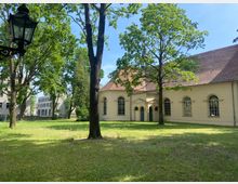 Historisches Kirchengebäude mit gelber Fassade, großen Rundbogenfenstern und einem roten Ziegeldach, umgeben von einer Rasenfläche und hohen Bäumen; links im Vordergrund eine schwarze Laterne. Im Hintergrund sind moderne Gebäude zu erkennen.