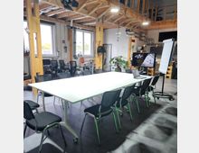 Moderner Besprechungsraum mit einem großen weißen Tisch, umgeben von schwarzen Stühlen. Im Hintergrund sind weitere Arbeitsplätze, Pflanzen, Holzbalken sowie eine helle Decke mit sichtbarer Technik zu sehen, ergänzt durch ein Display und ein Flipchart.