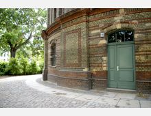 Backsteinfassade eines historischen Gebäudes mit Rundbogenfenstern und grüner Tür, umgeben von einem gepflasterten Weg und Bäumen im Hintergrund.