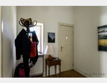 Ein schmaler Eingangsbereich mit einer weißen Tür, einem kleinen Holztisch mit einer Tischlampe und einer Garderobe, auf der Jacken und Hüte hängen. Rechts und links befinden sich gerahmte Bilder an den weißen Wänden.