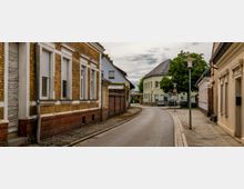 Eine schmale, gepflasterte Straße in einer ruhigen Wohngegend, gesäumt von Häusern mit Klinkerfassaden und weißen Fensterrahmen; im Hintergrund Kurve mit weiteren Gebäuden, darunter ein größeres, hellgelbes Haus. Straßenlaternen und vereinzelte Autos an den Seiten prägen die Szene, umgeben von Bäumen und Gehwegen.