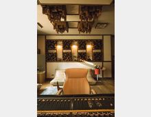 Tonstudio mit einem Mischpult im Vordergrund und einem beigen Ledersessel in der Mitte. Im Hintergrund befinden sich eine beige Couch, schalldämpfende Holzpaneele an den Wänden und dekorative Holzmodule an der Decke.